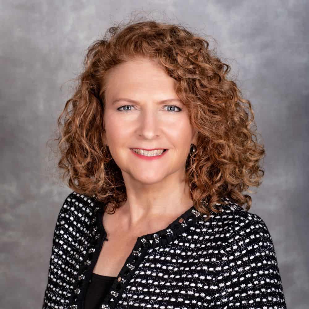 Confident woman with curly red hair in professional attire, smiling against a neutral background, showcasing leadership and expertise.