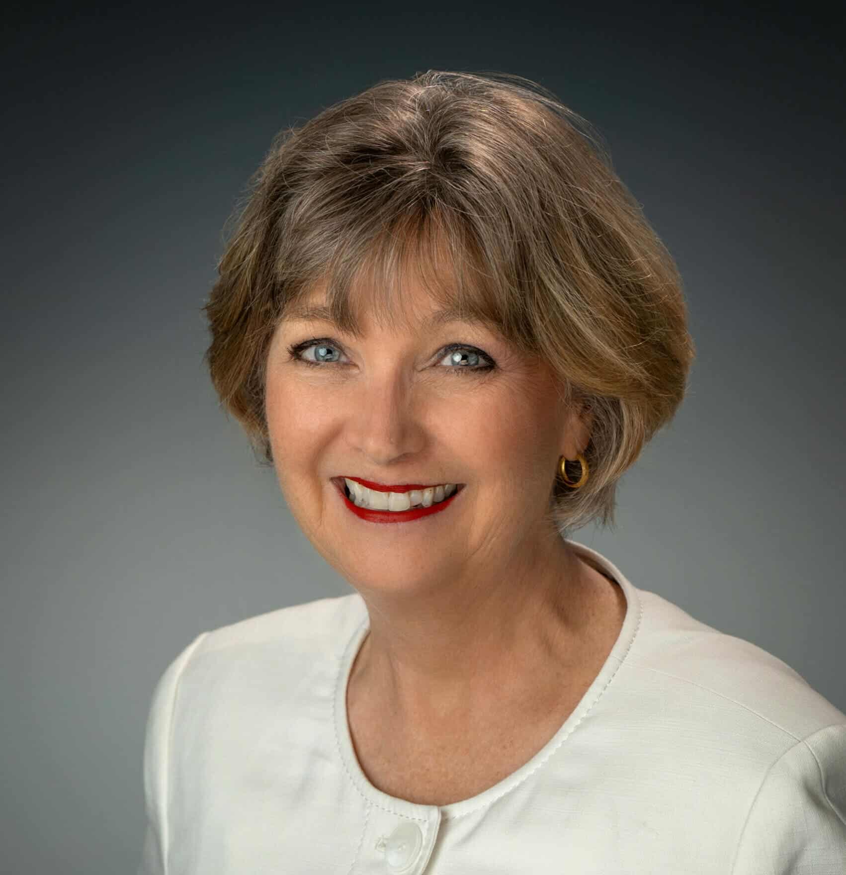 Portrait of a professional woman with light brown hair and blue eyes, smiling confidently in a white blazer.