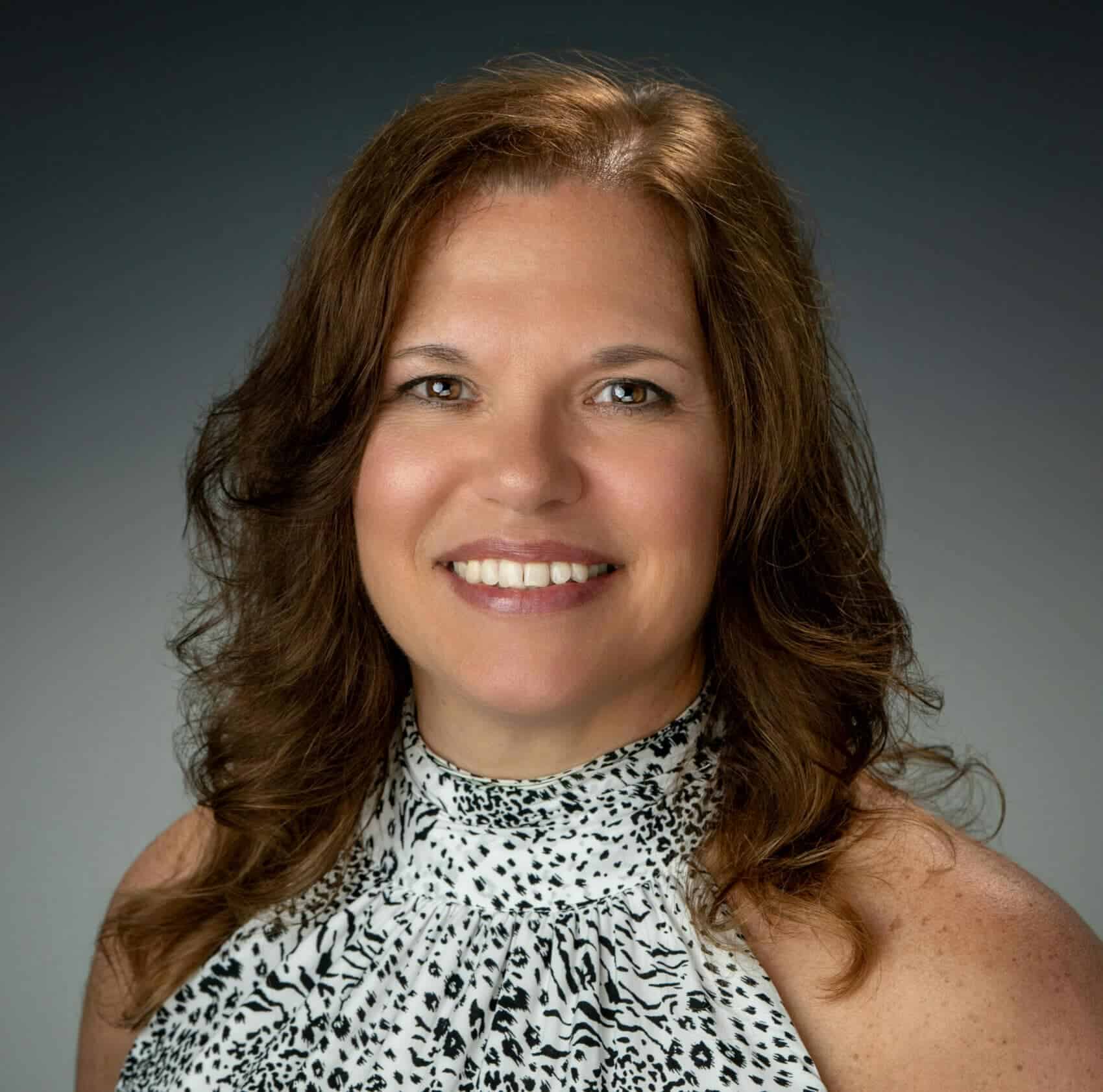 Friendly woman with shoulder-length wavy brown hair smiling against a dark gradient background for professional headshot.