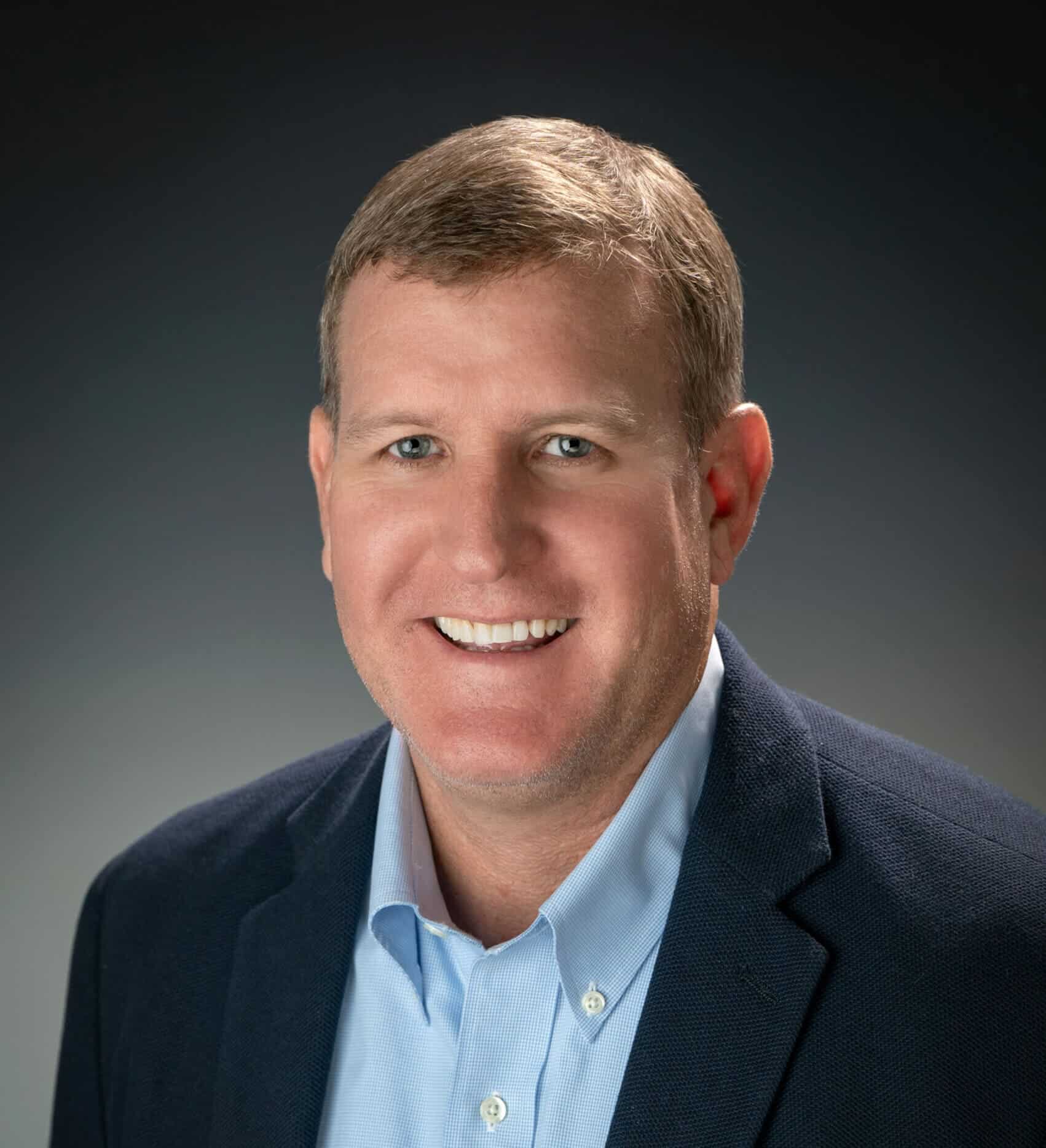 Image of a professional man smiling, wearing a navy blazer and light blue dress shirt, professional headshot, clean background.