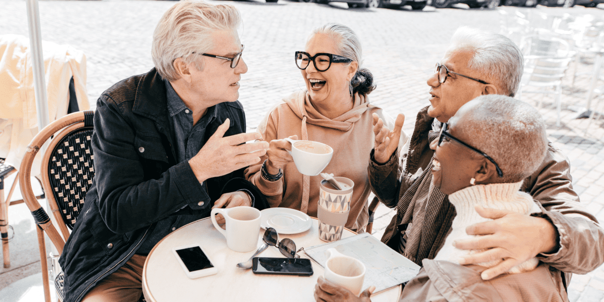 Gathering of diverse seniors enjoying coffee and conversation at an outdoor cafe in a sunny, urban setting, emphasizing friendship and social engagement among elderly people.