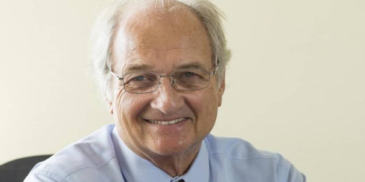 Photo of an elderly businessman wearing a light blue shirt and red tie, smiling confidently at the office.