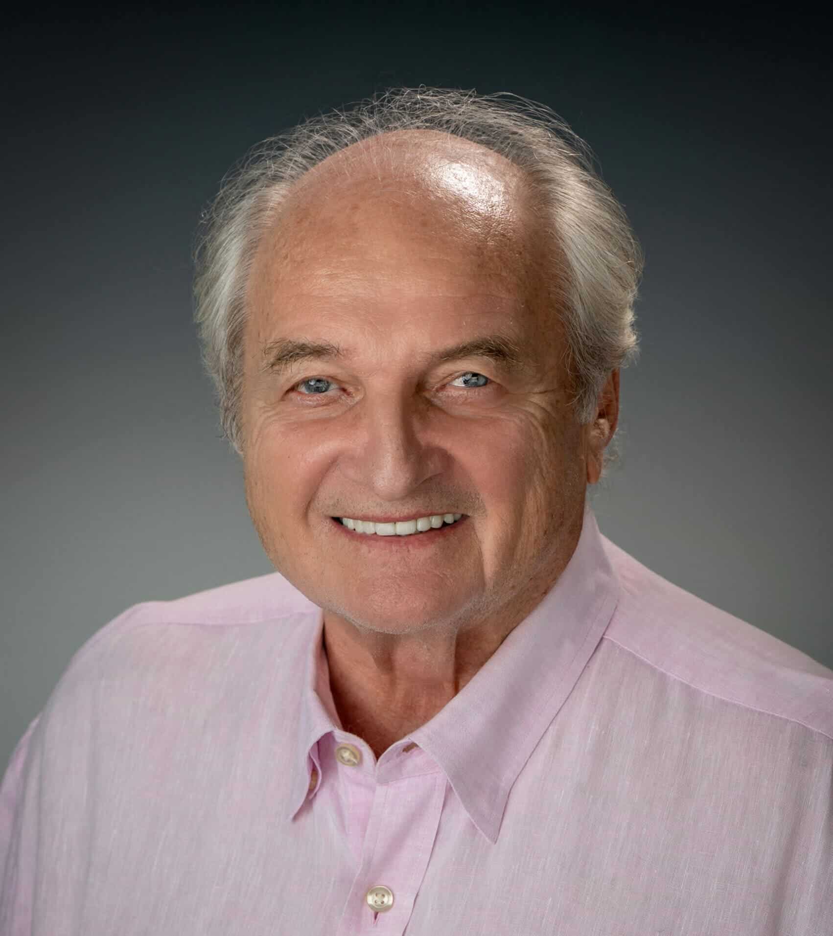 Elderly man with gray hair and blue eyes wearing a light pink shirt, smiling, business headshot, friendly demeanor.