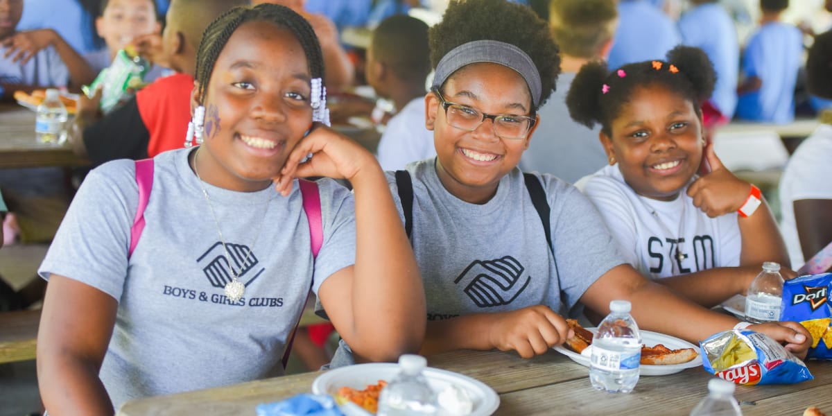 Smiling children at a summer camp eating pizza and snacks, wearing Boys & Girls Clubs t-shirts, diversity and friendship, cheerful youth engagement, vibrant community event photo. For Boys & Girls Clubs of Metropolitan Baltimore, a Carter partner.