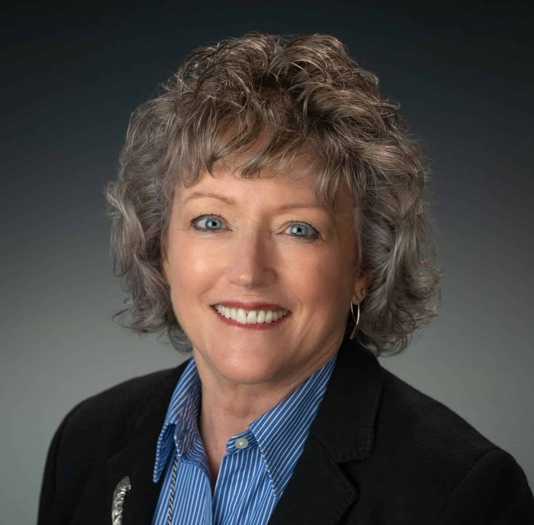 Friendly professional woman with curly gray hair smiling, wearing a blazer and blue striped shirt, headshot for business profile.