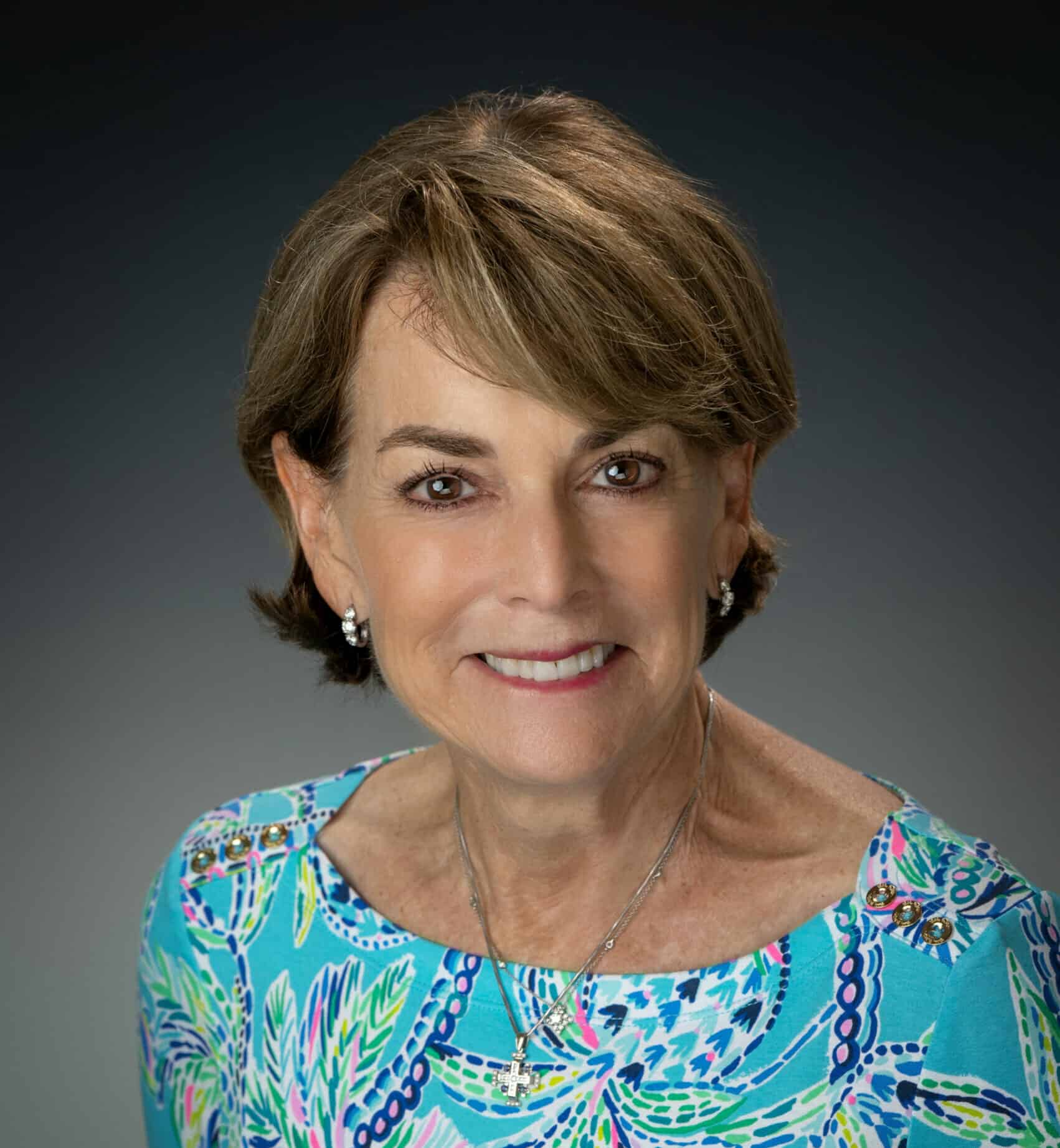 Professional portrait of a mature woman with short brown hair, wearing a vibrant blue patterned blouse, jewelry, and a warm smile.