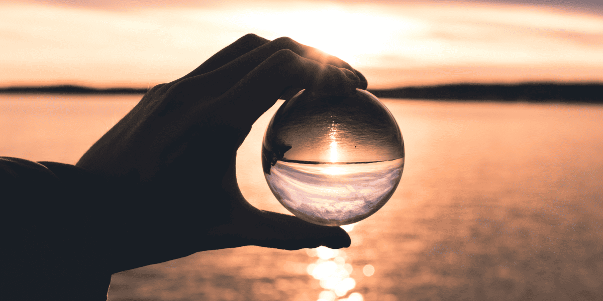 A hand holding a crystal ball reflecting a sunset over a calm ocean at dusk, showcasing serenity and natural beauty.