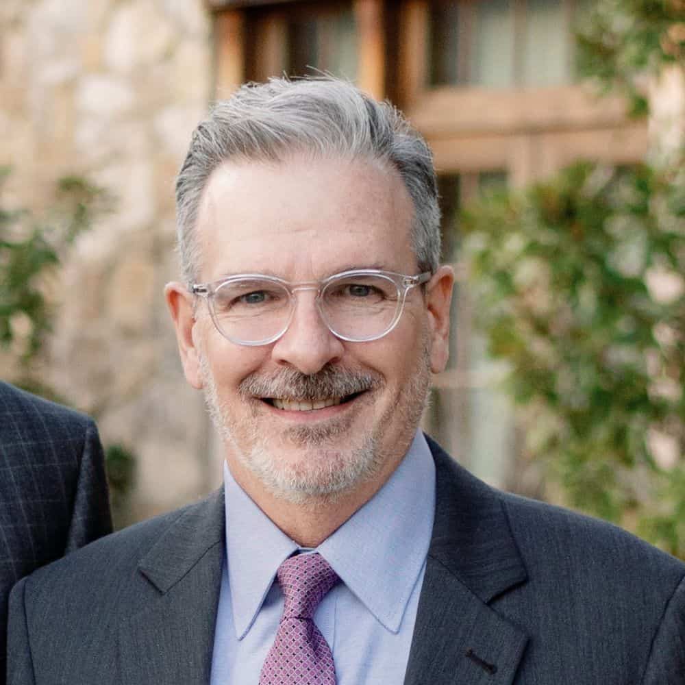 Professional man in business suit with glasses smiling outdoors, corporate headshot, executive portrait, smiling professional, business leader photo, modern executive headshot, business success, leadership, professional appearance, confident businessman wearing formal attire.