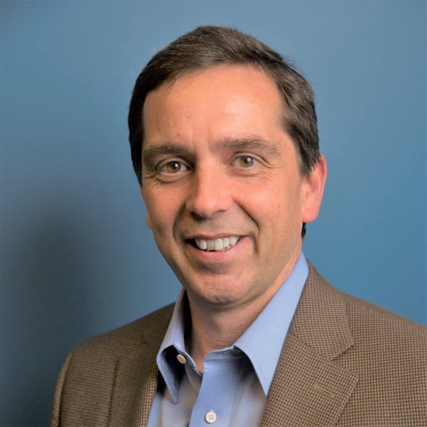 Experienced professional in business leadership with a confident smile, wearing a brown blazer and blue shirt, standing against a solid blue background. Perfect for corporate or executive profile images.