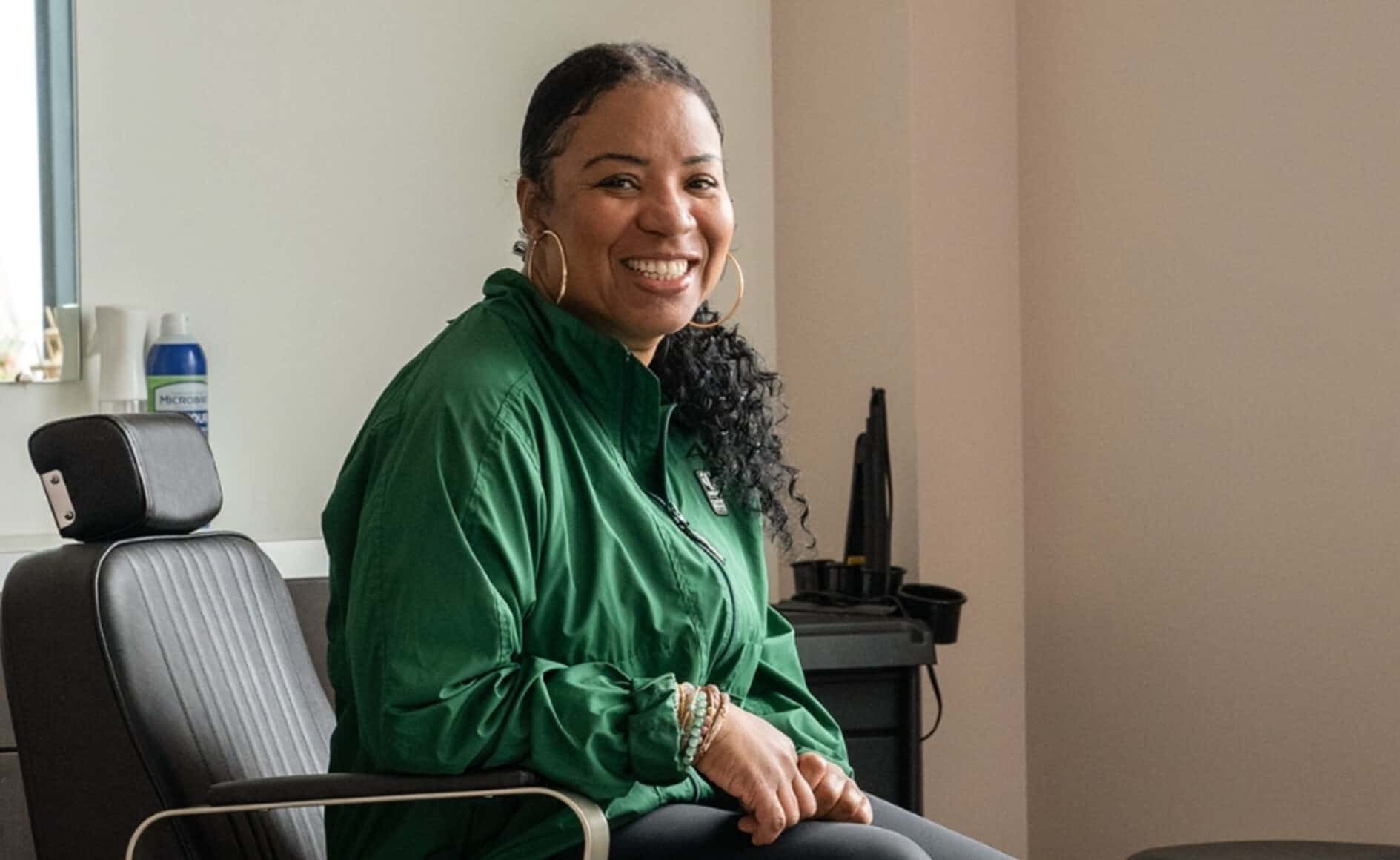 Smiling woman in a green jacket sitting in an office chair, professional and friendly atmosphere, corporate setting, upbeat business environment.