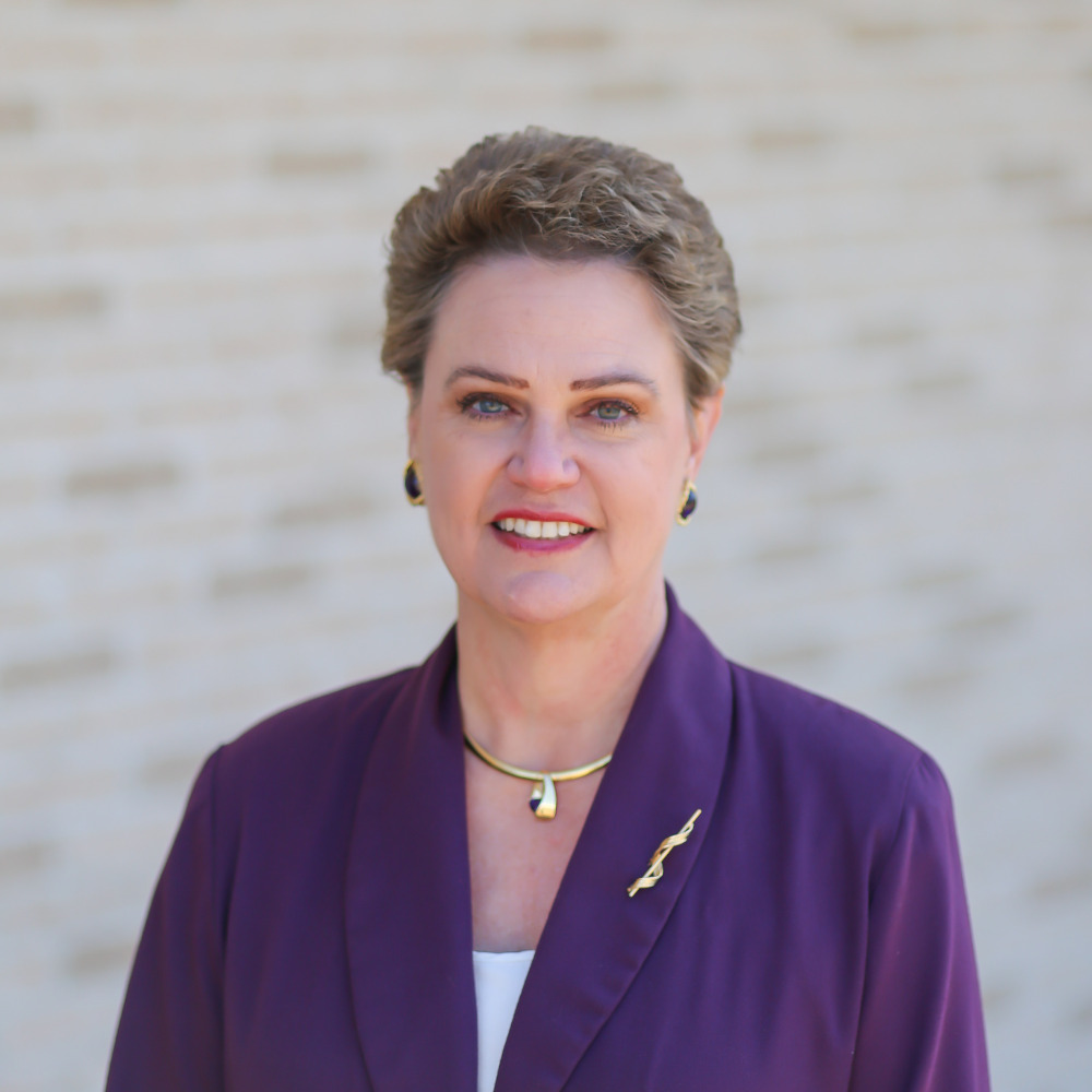 A professional woman with short hair wearing a purple blazer and jewelry, smiling outdoors.