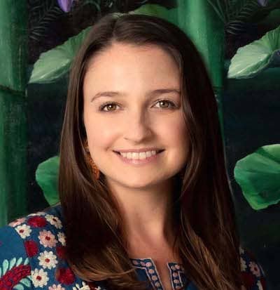 Smiling woman in vibrant floral blouse with lush green tropical background.