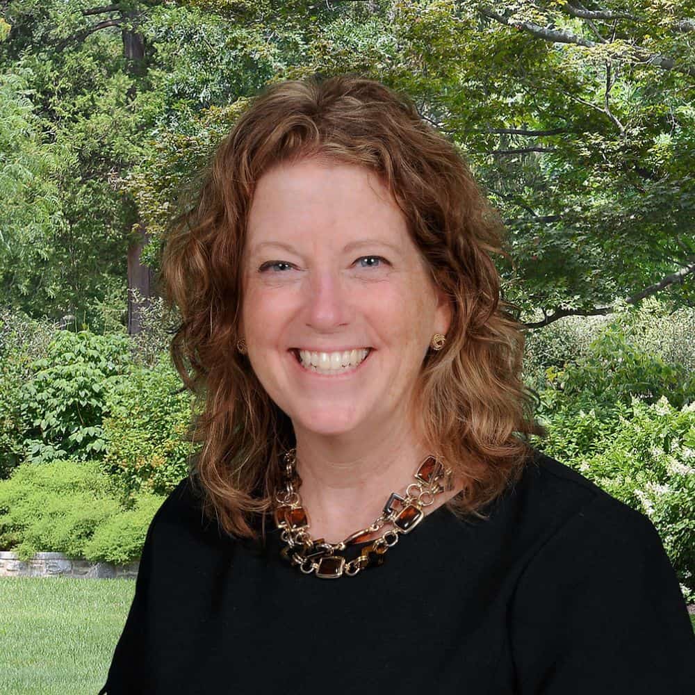 Vibrant portrait of a smiling woman with curly red hair standing outdoors in a lush green garden, showcasing professional headshot styling for business and personal branding.