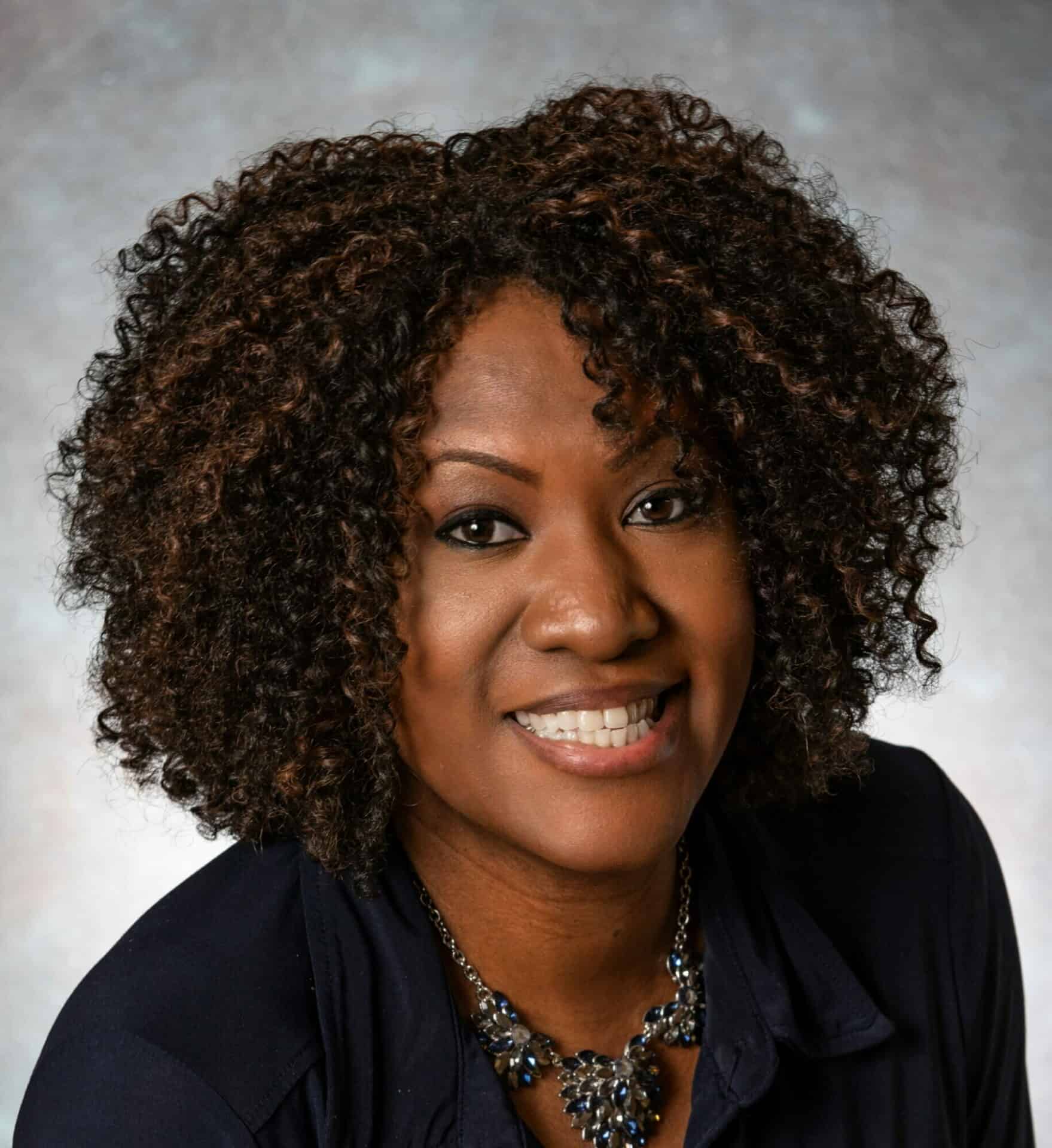 Professional headshot of an African American woman with natural curly hairstyle, smiling and wearing a black blazer and necklace, on a neutral backdrop.