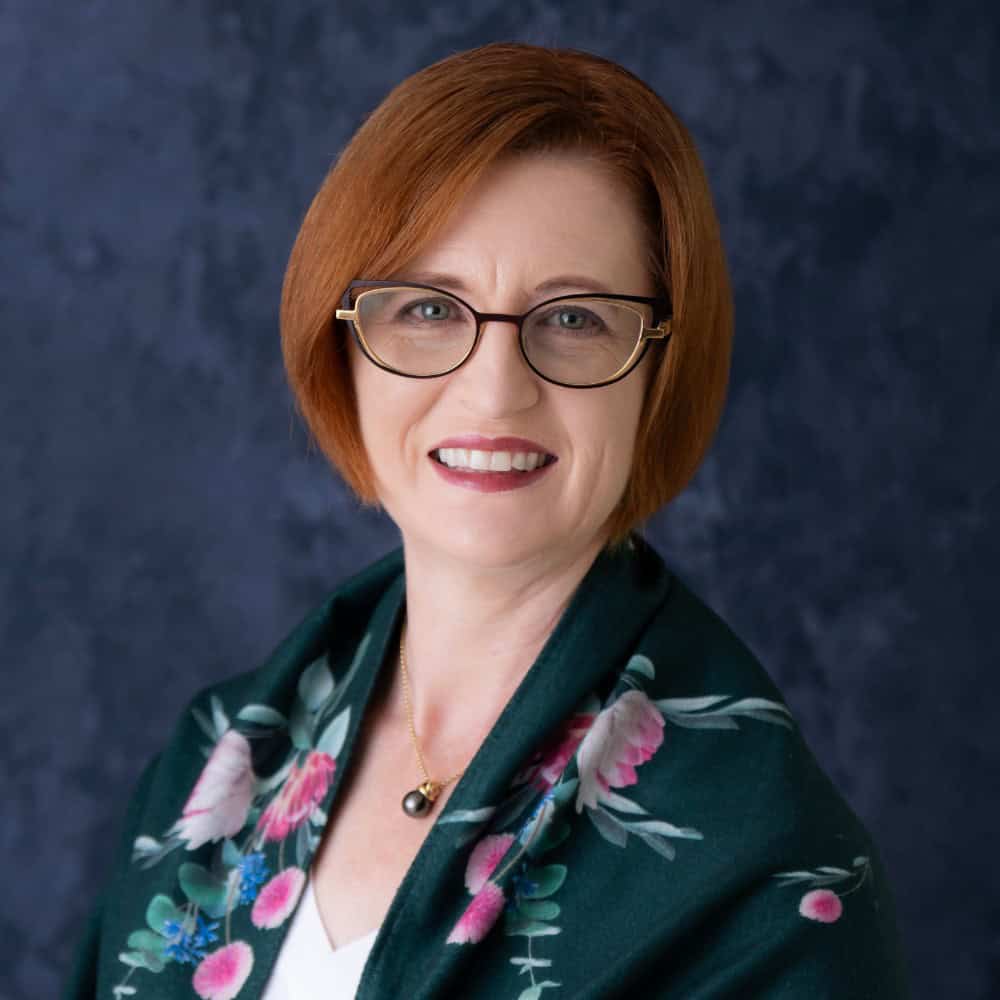 Elegant red-haired woman wearing glasses and floral blazer, professional headshot for corporate or business profile.