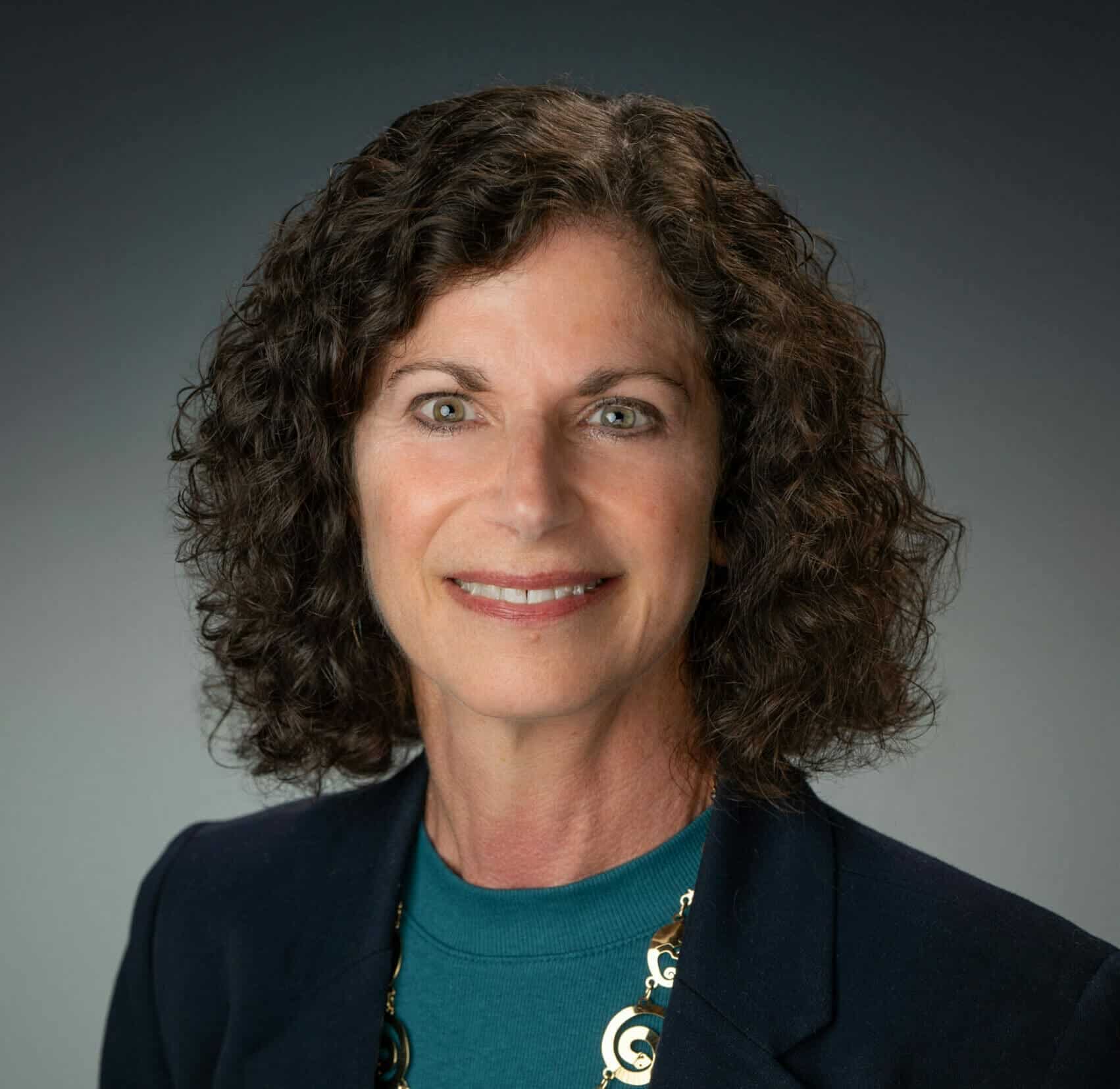 Professional woman with curly brown hair, wearing a dark blazer and teal top, smiling confidently for a headshot.