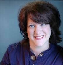 A professional headshot of smiling woman with shoulder-length dark hair and hoop earrings, wearing a dark top, against a neutral background.