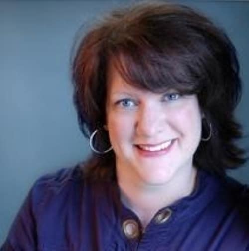 A professional headshot of smiling woman with shoulder-length dark hair and hoop earrings, wearing a dark top, against a neutral background.