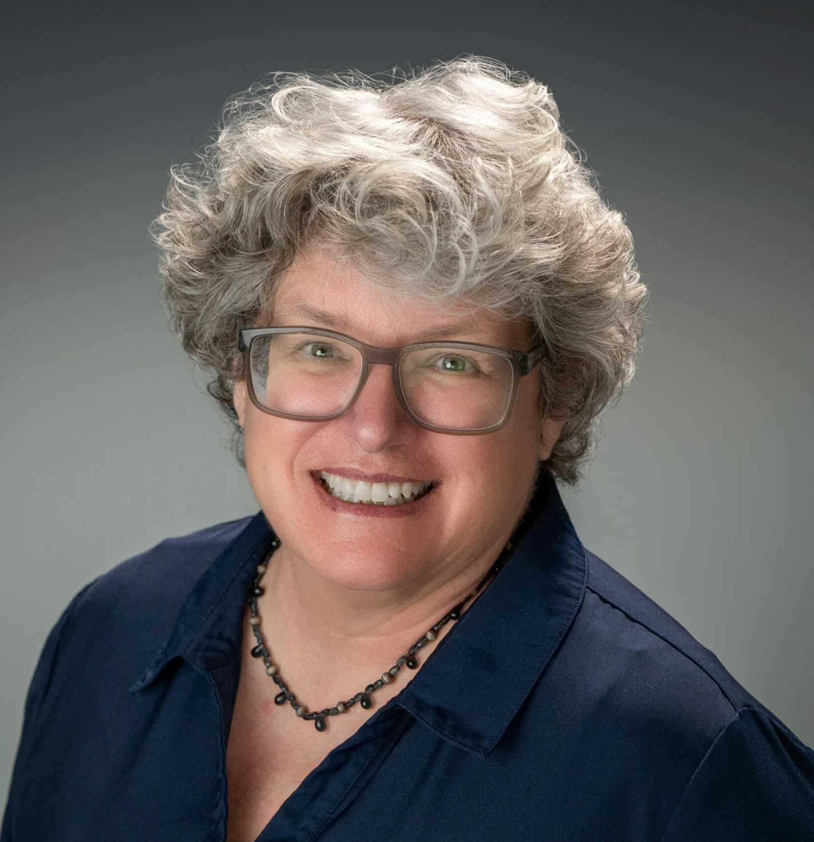Smiling woman with curly gray hair and glasses in professional portrait.