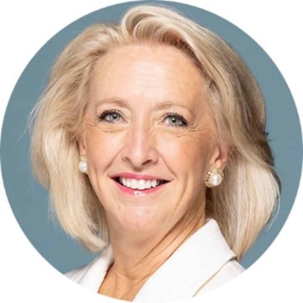 Bright professional woman with blonde hair, blue eyes, and pearl earrings, smiling in a white blazer for a corporate or business profile picture.