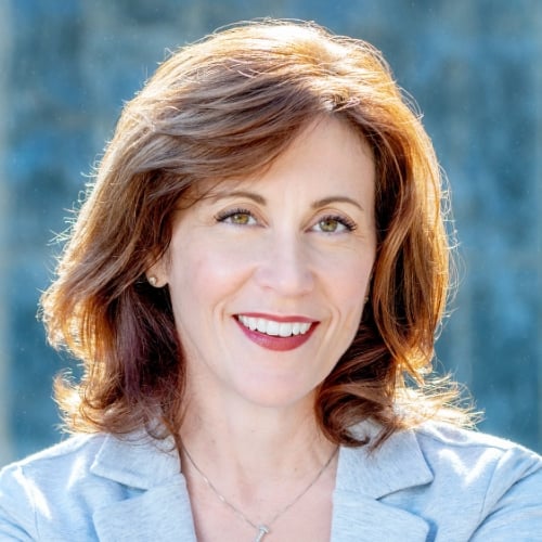 Smiling woman with red hair wearing a light gray blazer outdoors, representing confidence and professionalism.