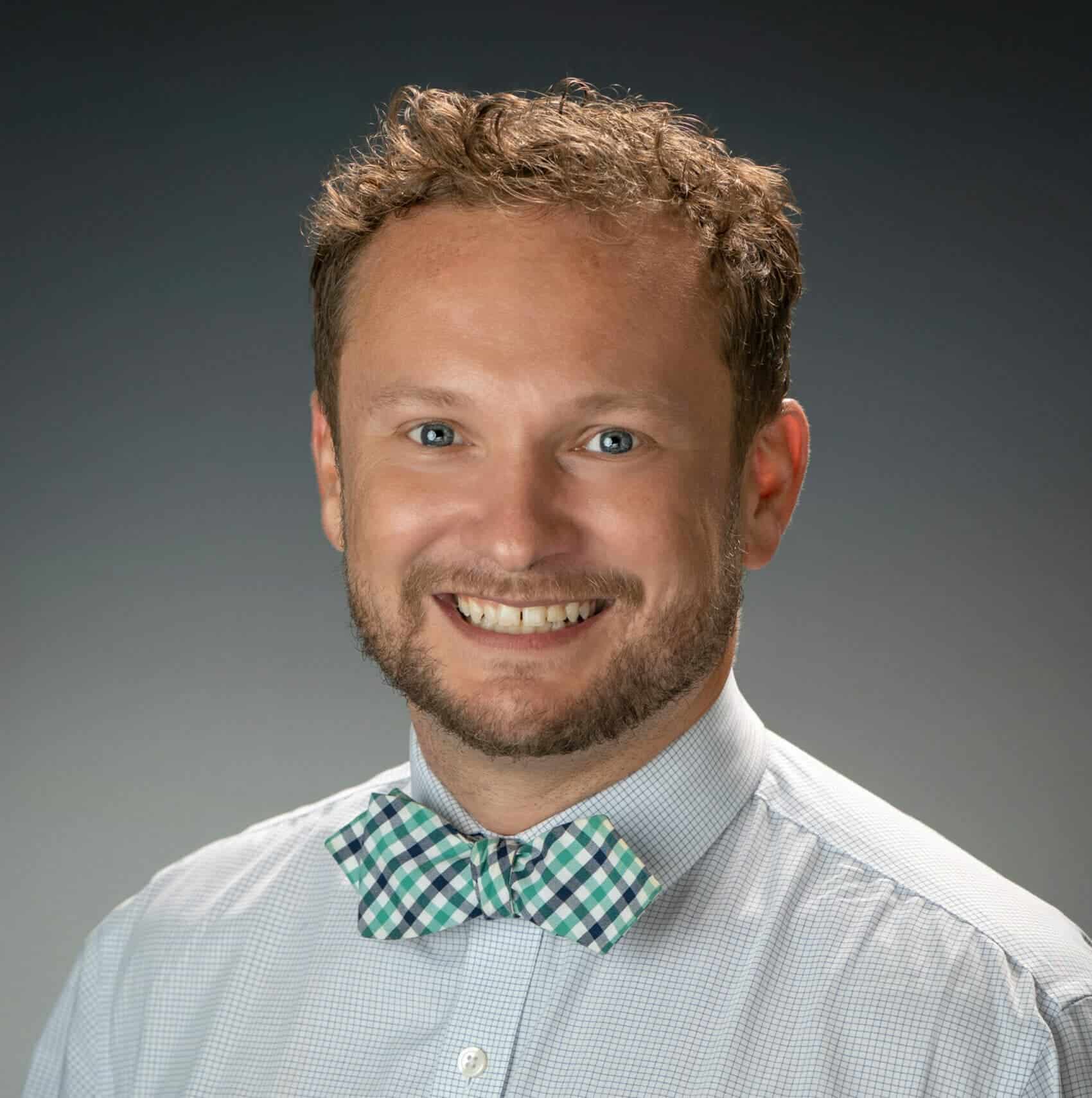 Happy young man with curly hair and beard in a professional headshot, wearing a checkered bow tie and dress shirt, smiling confidently.