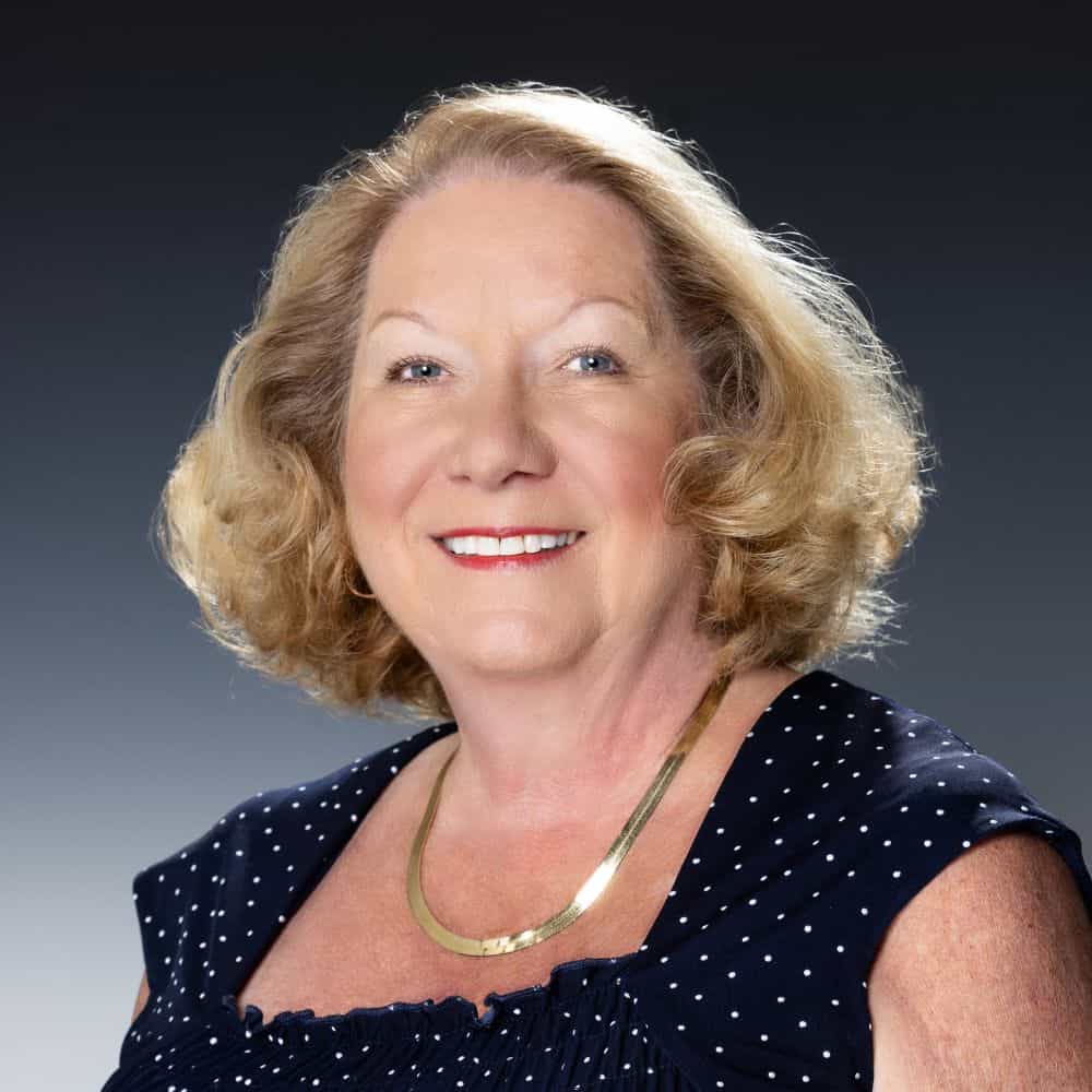 Senior woman smiling in a professional portrait, wearing a navy polka dot top and gold necklace, with styled blonde hair and a confident demeanor.