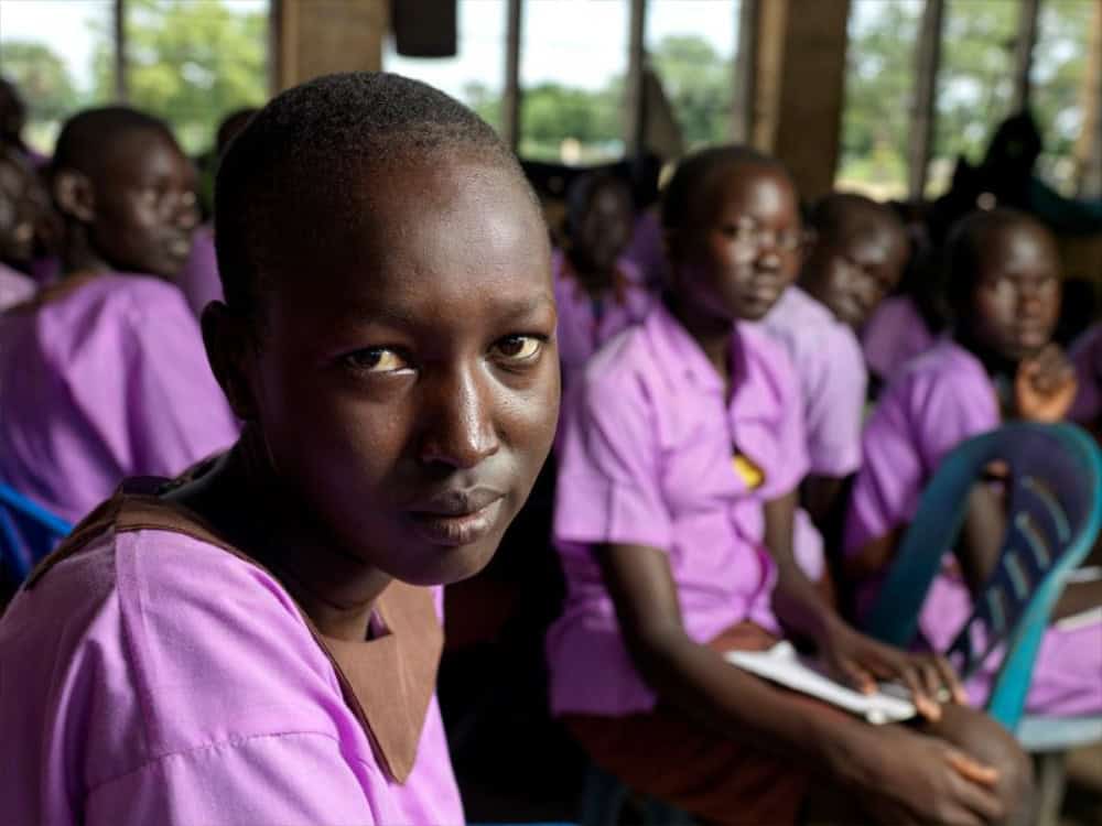Young African girl in purple school uniform sitting in classroom with other students, focused on learning and education, classroom setting, community, and youth empowerment for Plan International USA, a Carter partner.