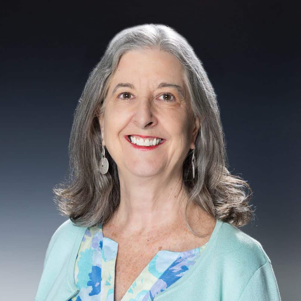 Portrait of a smiling senior woman with gray hair, wearing a light blue top, on a dark gradient background for healthcare and aging-related content.