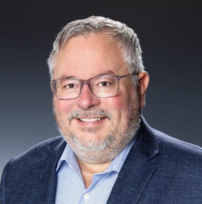Warm professional headshot of a smiling man with glasses, gray hair, wearing a blue blazer and shirt.