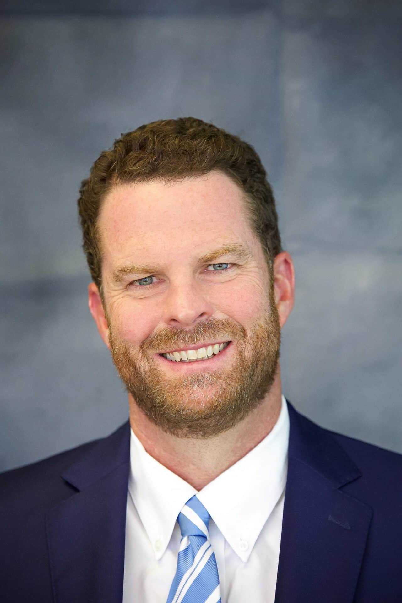 Professional headshot of a smiling man in a navy suit and light blue striped tie, confident.