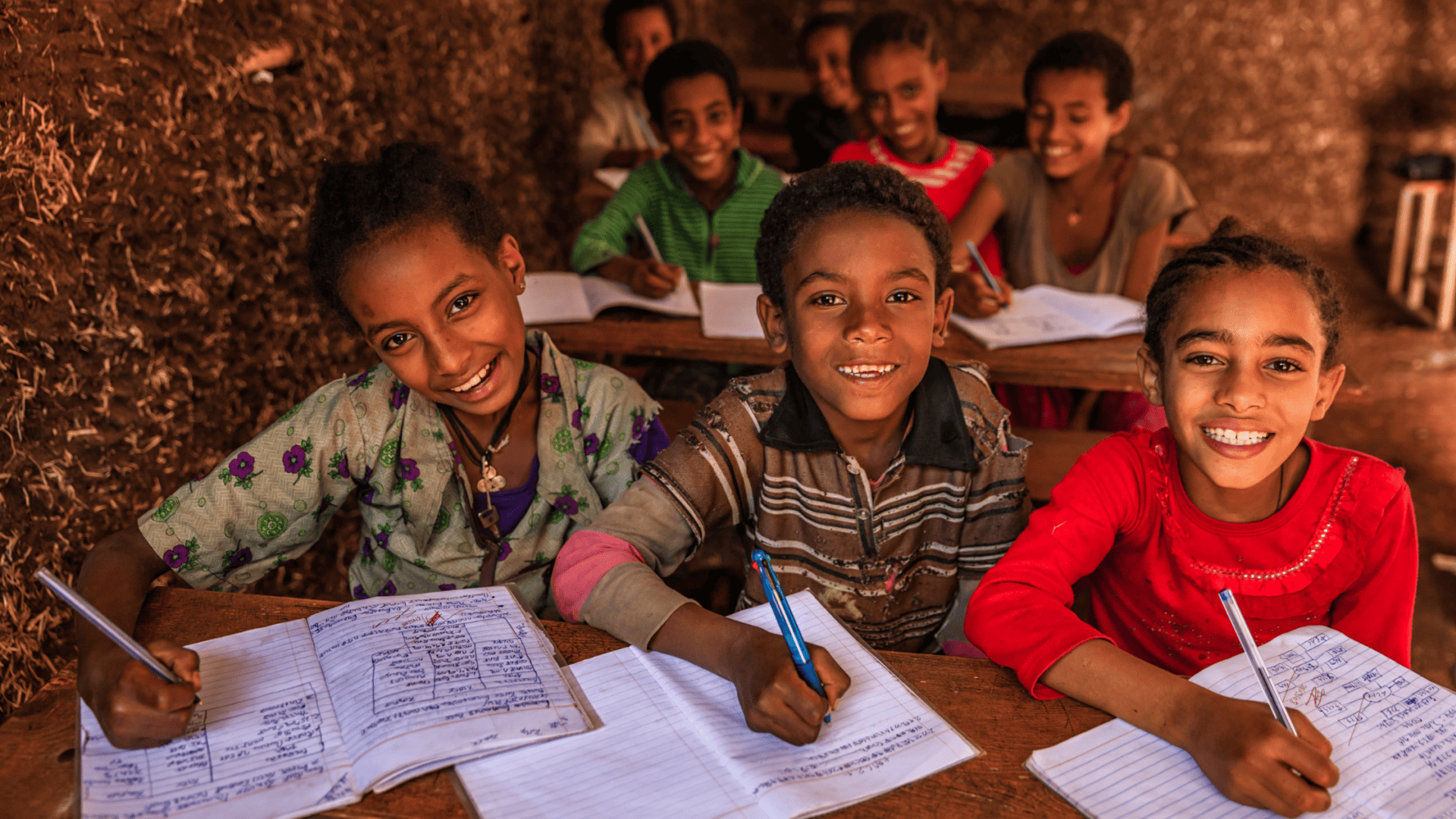 Smiling children in a classroom, studying and writing in notebooks, outdoors setting, future leaders, educational success, joyful learning experience.