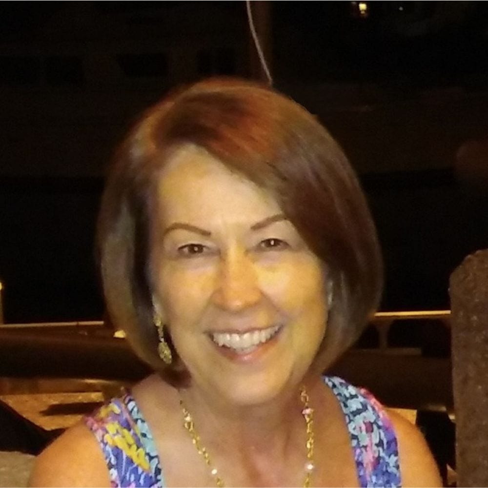 Bright smiling woman with short brown hair at an outdoor evening event, wearing colorful sleeveless dress, jewelry, and earrings, in a warm, festive atmosphere.