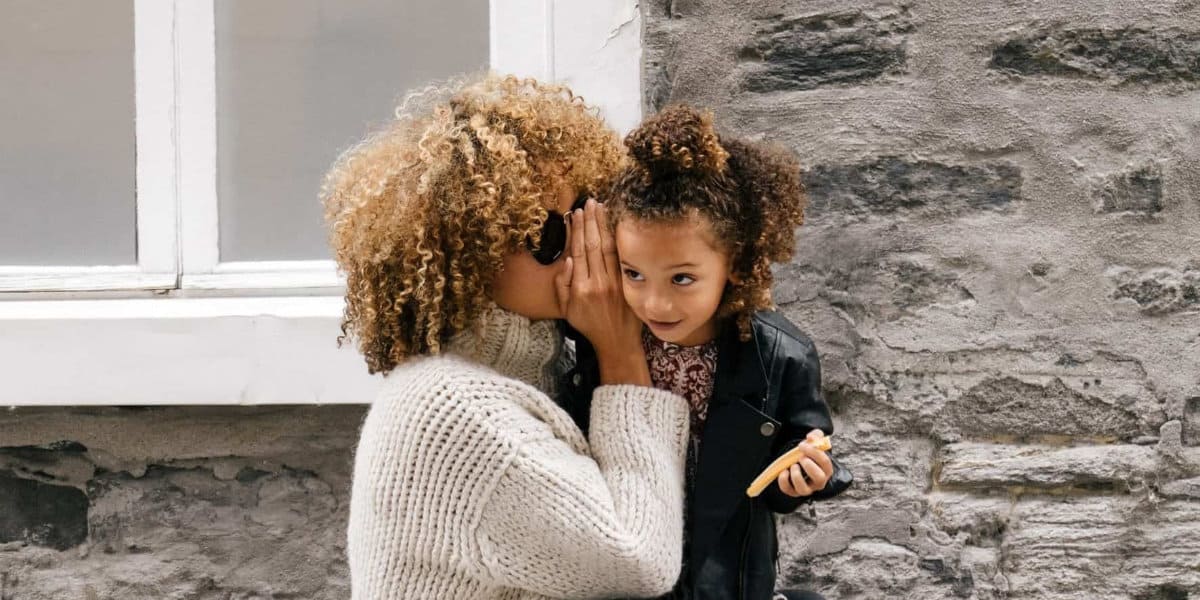 Whispering to a young girl with curly hair, outdoor city scene, mother and daughter sharing secrets, casual fashion, stone wall background, warm family moment, close-up of communication, candid portrait.
