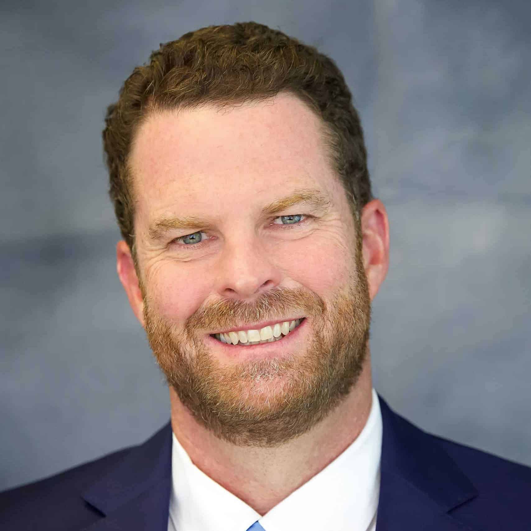 Bright smiling man with blue eyes, red beard, and dressed in a navy suit and white shirt, professional portrait for corporate or business profile.