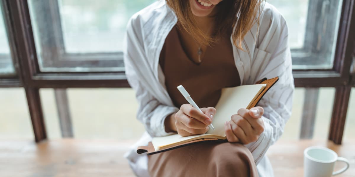 Thoughtful young woman writing in a notebook by a window, capturing ideas for creative journaling and work productivity.